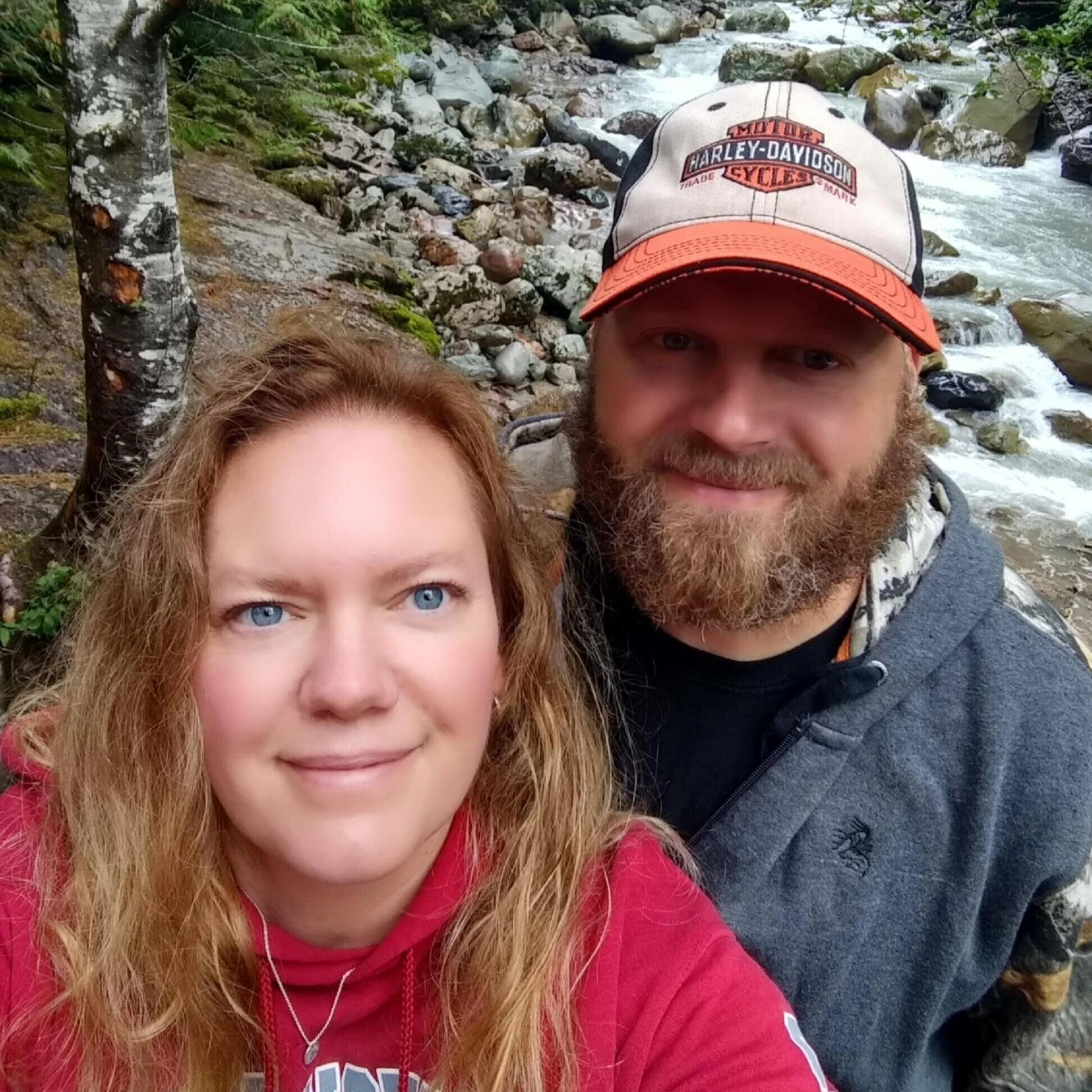 Couple taking a selfie by a river in a lush forest, showcasing the natural beauty of the outdoors, with rocks and flowing water in the background. The woman has long, wavy hair and is wearing a red hoodie, while the man has a beard and is wearing a Harley-Davidson cap.